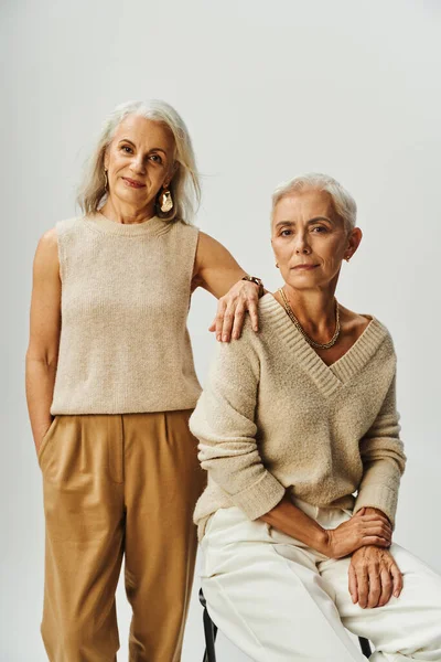 Elegante Seniorin sitzt neben lächelnder Freundin und blickt in die Kamera auf Grau, Mode und Stil — Stockfoto