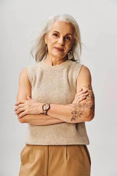 Modische und tätowierte Seniorin posiert mit verschränkten Armen und blickt in die Kamera auf grau — Stockfoto