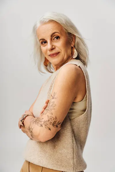Señora mayor tatuada y de moda en el oeste de punto posando con los brazos cruzados, sonriendo a la cámara en gris - foto de stock