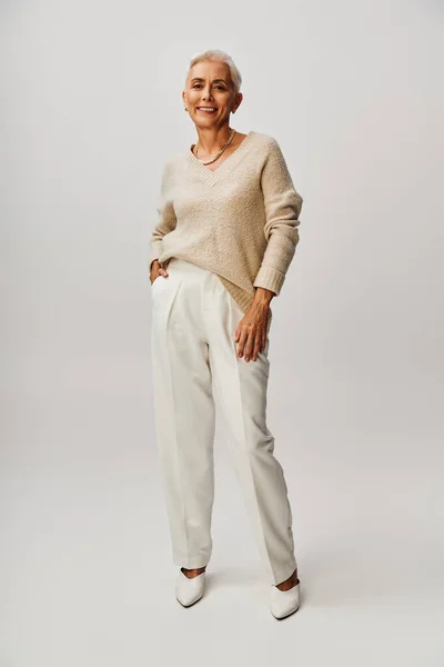 Donna matura alla moda in maglione annodato e pantaloni bianchi in posa con mano in tasca su grigio, lunghezza intera — Foto stock
