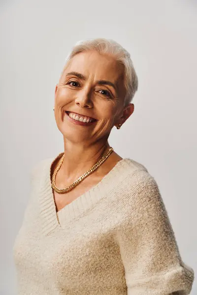 Cheerful senior model with makeup, short silver hair and golden necklace on grey backdrop, portrait — Stock Photo