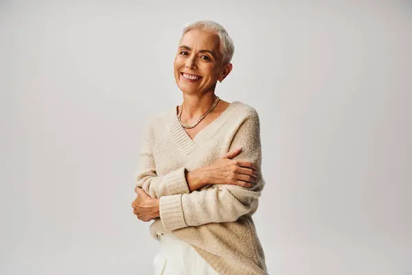 Joyful senior model in soft knitted jumper posing with folded arms and smiling at camera on grey — Stock Photo
