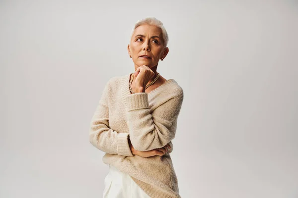 Мрійлива старша жінка в модному повсякденному вбранні тримає руку біля підборіддя і дивиться на сірий — стокове фото