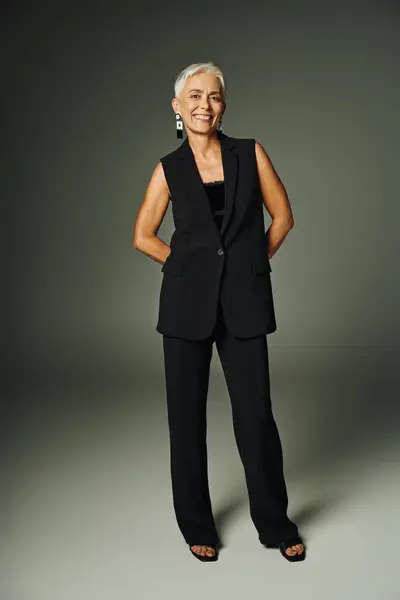Fashionable senior woman with radiant smile posing in black elegant attire on grey, full length — Stock Photo