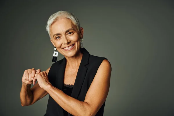 Happy mature lady in stylish earrings and black attire smiling at camera on grey, elegance and charm — Stock Photo