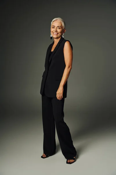 Full length of smiling senior model in black classic clothes looking at camera and standing on grey — Stock Photo