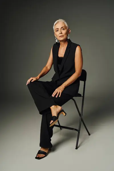 Senhora sênior na moda em traje elegante preto sentado na cadeira em cinza, moda clássica e idade — Fotografia de Stock