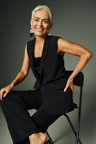 Envejecimiento con gracia, mujer mayor sonriente en ropa negra sentado en la silla con la mano en la cadera en gris - foto de stock