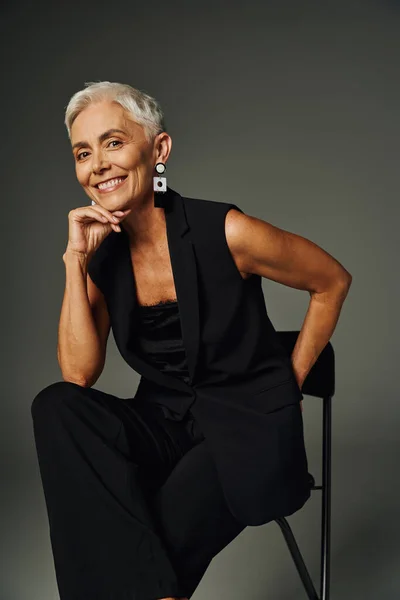 Позитивна, весела старша жінка в чорному вбранні сидить на стільці з рукою позаду на сірому — стокове фото
