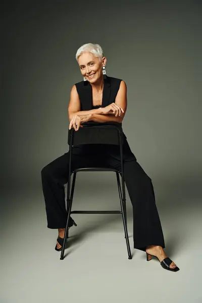 Full length of joyful and graceful mature lady in black sitting on chair and smiling on grey — Stock Photo