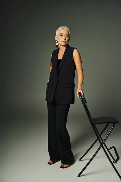 Full length of confident senior woman in black attire posing with chair on grey, classic fashion — Stock Photo