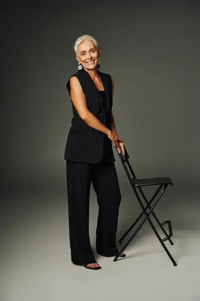 Longitud completa de señora mayor alegre en traje clásico negro posando con silla en gris, elegancia - foto de stock