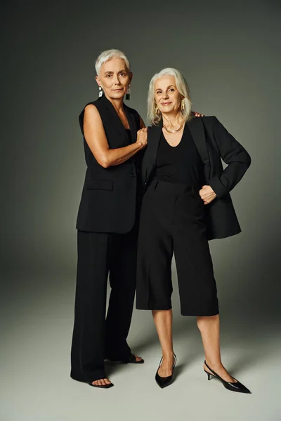 Усміхнена старша леді в чорному одязі позує з рукою на стегні поруч зі стильною подругою-жінкою на сірому — стокове фото