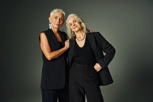 Щаслива старша модель в чорному вбранні позує з рукою на стегні біля елегантної подруги на сірому — стокове фото