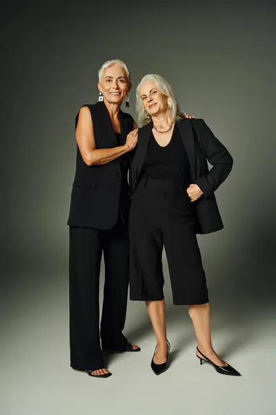 Alegres seniores amigos do sexo feminino em trajes elegantes pretos olhando para a câmera em cinza, envelhecimento da moda — Fotografia de Stock
