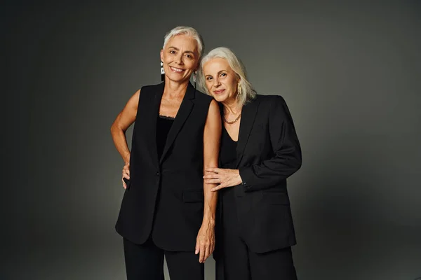 Pleased senior models in black fashionable clothes looking at camera on grey, charm and elegance — Stock Photo
