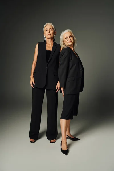 Повна довжина витончених літніх жінок в чорному стильному одязі, що стоїть на сірому, класичному стилі — стокове фото