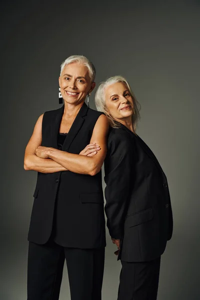 Joyful mature woman with folded arms near smiling female friend on grey, fashionable seniors — Stock Photo
