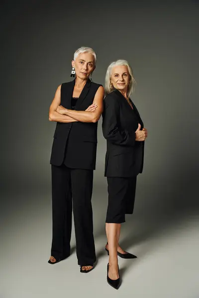 Trendy senior lady in black attire posing with folded arms near female friend on grey, elegance — Stock Photo