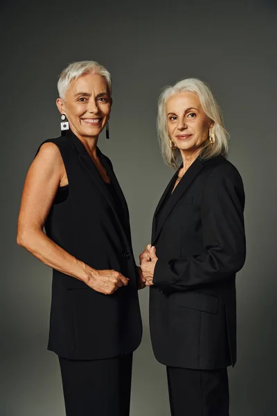 Elegant senior female friends in black stylish clothes smiling at camera on grey, vanity fair style — Stock Photo