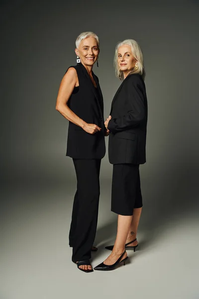 Positive mature women  in black elegant wear smiling at camera on grey, senior fashion, full length — Stock Photo