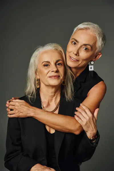 Fashionable senior lady in black attire embracing trendy female friend on grey, elegant aging — Stock Photo