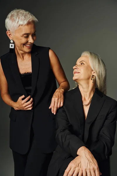 Alegres e na moda amigos do sexo feminino ao longo da vida em roupas pretas olhando uns para os outros em cinza, positividade — Fotografia de Stock