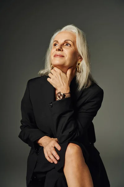 Femme mature rêveuse et souriante en noir élégant porter assis et regardant loin sur gris, élégance — Photo de stock