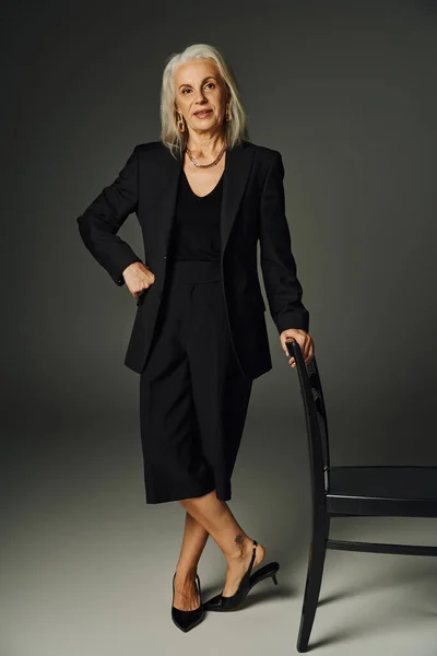 Full length of senior model in black attire posing with hand on hip near chair on grey, elegance — Stock Photo