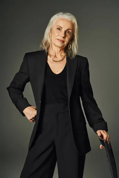 Élégant modèle senior en vêtements noirs et accessoires dorés près de la chaise avec la main sur la hanche sur gris — Photo de stock