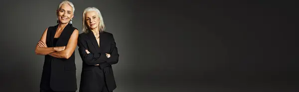 Felice senior fashionistas in nero abbigliamento classico in posa con le braccia piegate su sfondo grigio, banner — Foto stock