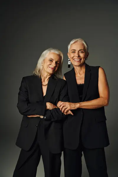 Joyeuses amies aînées en costume élégant noir souriant à la caméra tout en posant sur le gris — Photo de stock