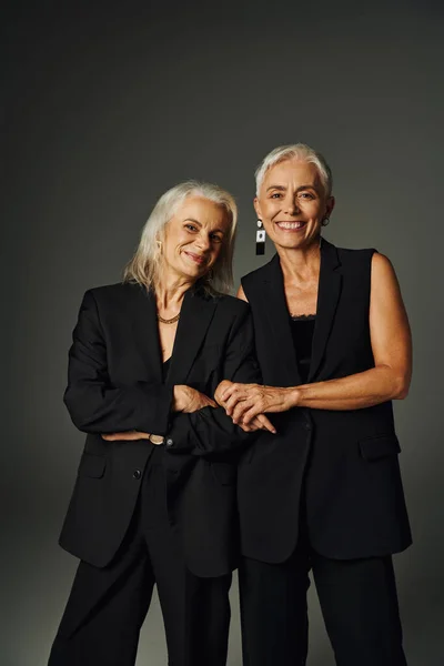 Joyeuses amies aînées en costume élégant noir souriant à la caméra tout en posant sur le gris — Photo de stock