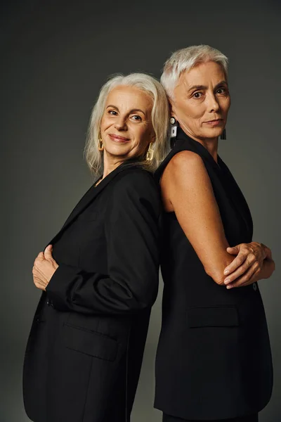Senior models in black classic clothes standing back to back and looking at camera on grey — Stock Photo
