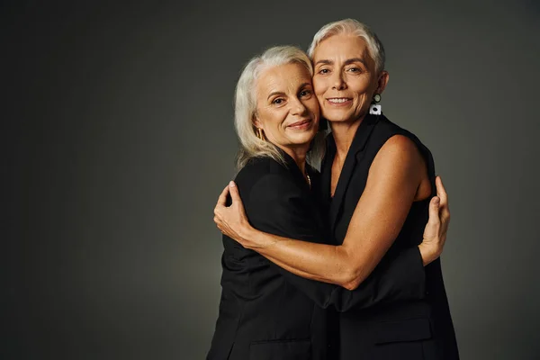 Amigos ao longo da vida, senhoras seniores alegres e elegantes em roupas pretas abraçando em pano de fundo cinza — Fotografia de Stock