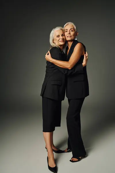 Повна довжина позитивних і модних старших моделей в чорному вбранні, що обіймають один одного на сірому тлі — стокове фото