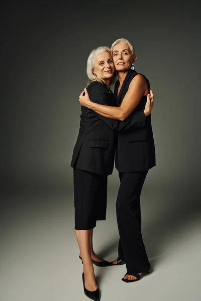 Full length of positive and trendy senior models in black attire hugging each other on grey backdrop — Stock Photo