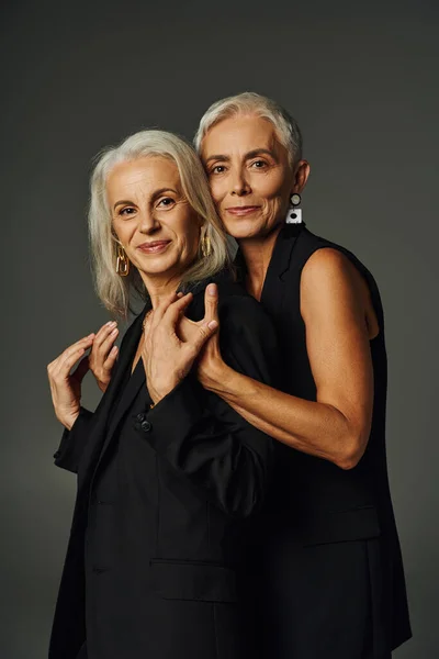 Elegant mature lady in black wear hugging shoulders of female friend and looking at camera on grey — Stock Photo