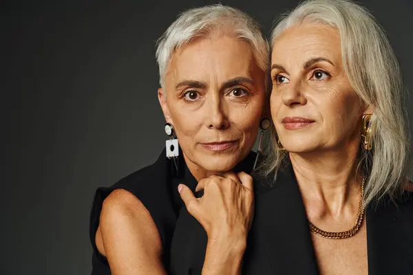 Mature model in black stylish clothes embracing shoulders of female friend on grey backdrop — Stock Photo