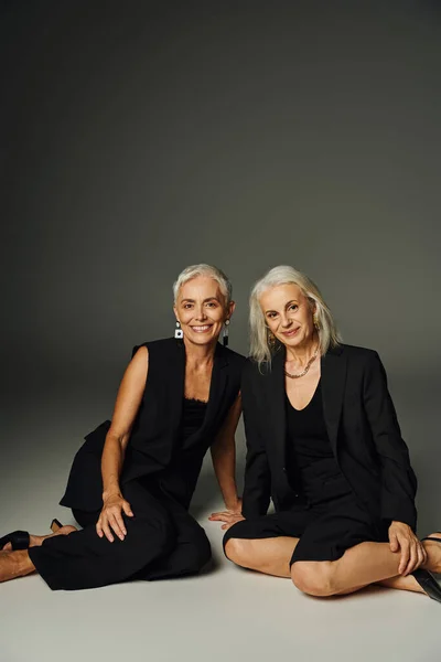 Dames matures à la mode en vêtements noirs assis et souriant à la caméra sur gris, mode classique — Photo de stock