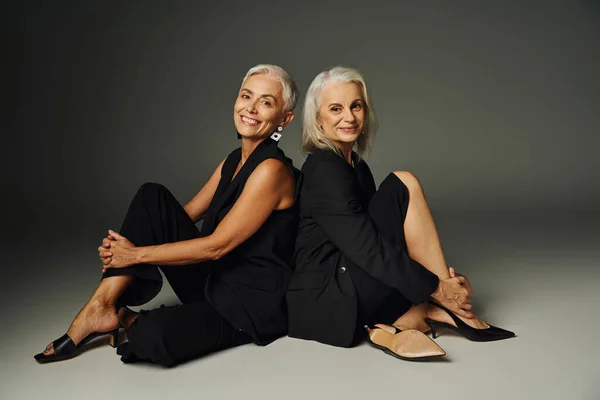 Mulheres seniores alegres e na moda em roupas pretas sentadas de costas para trás em pano de fundo cinza, elegância — Fotografia de Stock