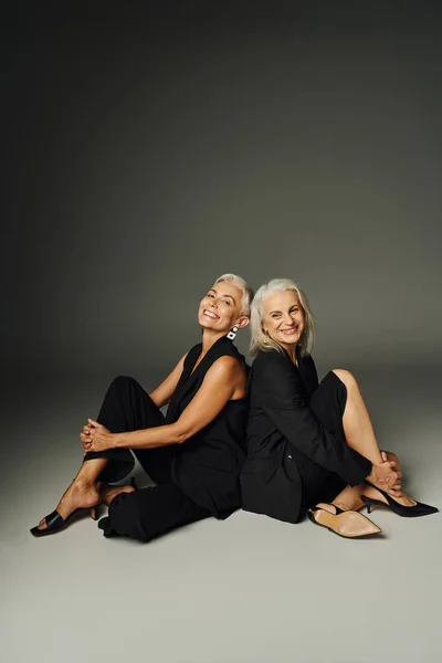 Alegre sénior amigas en traje negro sentado espalda con espalda en gris, envejecimiento de moda - foto de stock