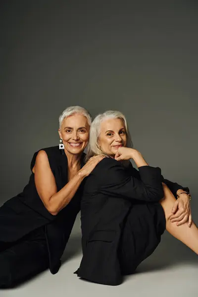 Amigos do sexo feminino seniores na moda em roupas pretas sentado e sorrindo para a câmera em cinza, positividade — Fotografia de Stock