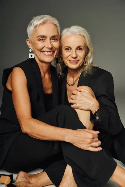 Delighted senior fashionistas in black clothes sitting and looking at camera on grey, friendship — Stock Photo