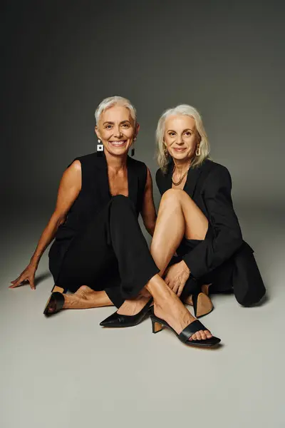 Positif senior trendsetters en tenue noire assis et souriant à la caméra sur gris, mode classique — Photo de stock