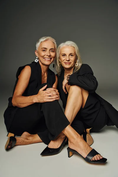 Joyeuses mannequins seniors en costume élégant noir assis regardant la caméra tout en étant assis sur le gris — Photo de stock