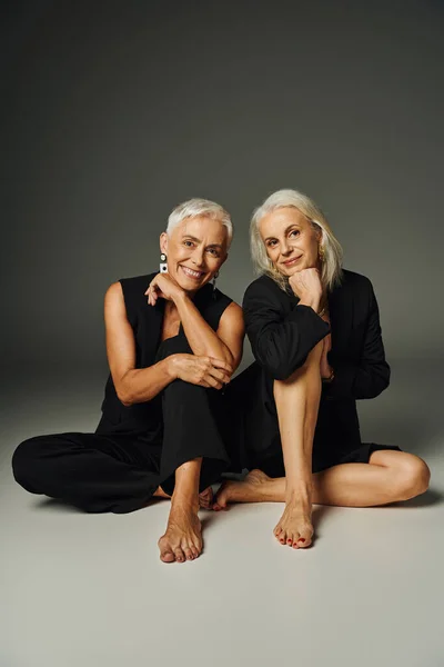 Cheerful barefoot senior models in black fashionable clothes sitting and looking at camera on grey — Stock Photo