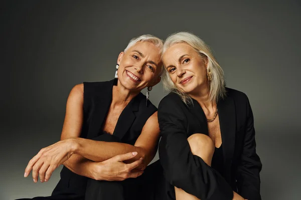 Senior female friends in black fashionable attire smiling at camera while posing on grey, positivity — Stock Photo