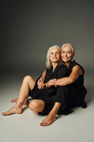 Barefoot senior female friends in black clothes sitting and looking at camera on grey, age positive — Stock Photo