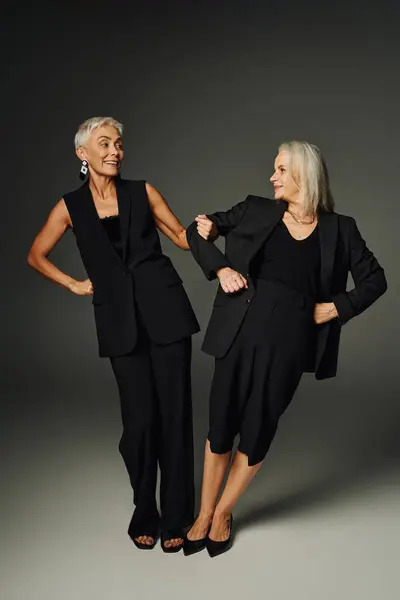 Senior female friends in black trendy wear having fun and smiling at each other on grey, full length — Stock Photo